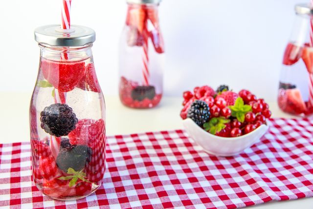 Colorful berry-infused water bottles arranged on a red and white checkered cloth. Fresh strawberries, blackberries, and raspberries combined in mason jars for a refreshing and healthy drink option. Nearby, a white bowl filled with a mix of fresh berries adds to the vibrant scene. Ideal for use in summer picnic or healthy eating related content, wellness blogs, or hydration-focused advertisements.
