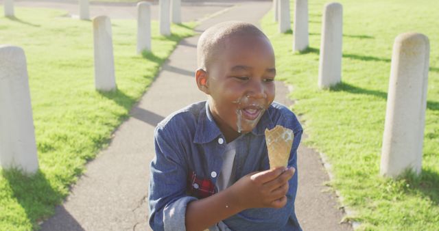 Joyful young boy enjoying ice cream in a sunny park. Dressed in a denim shirt, the child displays a messiness characteristic of childhood exuberance while relishing his treat. Perfect for advertisements on summer fun, childhood, family activities, and outdoor leisure. Suitable for adding heartwarming elements to promotional materials.