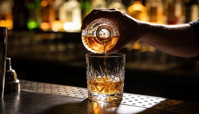 Bartender pouring amber whiskey into cut crystal lowball on bar rail with warm bokeh