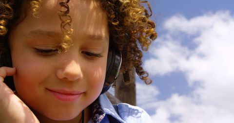 Biracial boy with curly hair enjoys music outdoors, sky in background