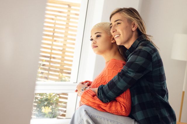 This image captures a multiracial lesbian couple embracing and looking thoughtfully out of a window at home. It can be used to represent themes of love, togetherness, and LGBTQ relationships. Ideal for articles, blogs, or advertisements focusing on relationships, diversity, and inclusivity.