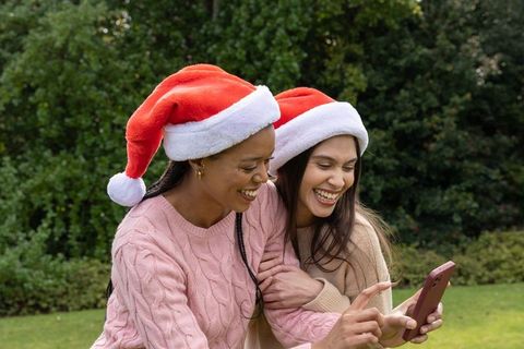 Diverse friends wearing santa hats and cozy knit sweaters laughing while looking at phone