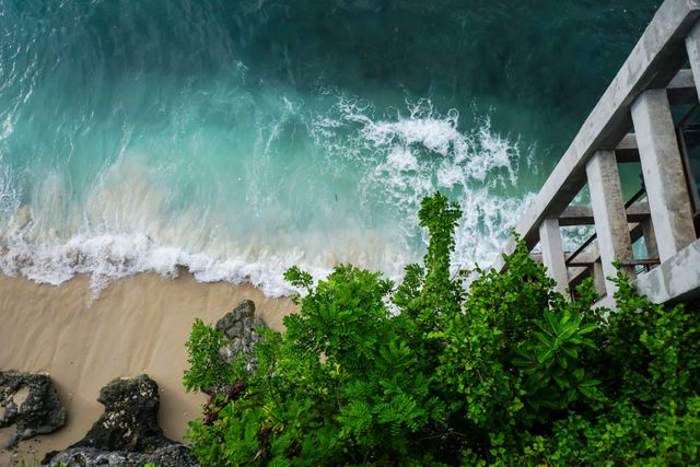 High angle view of a tropical beach with ocean waves crashing on white sand. Lush green plants add a touch of natural beauty. Ideal for travel brochures, summer vacation promotions, or websites focusing on exotic destinations and relaxation.