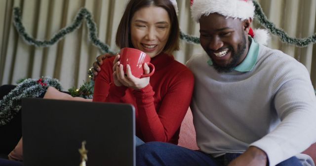 Image of happy diverse couple in santa hats smiling and making christmas laptop image call at home. Christmas, celebration, communication, happiness and inclusivity concept.