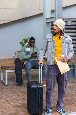 Urban friends waiting with suitcase and skateboard sharing street food and smartphone