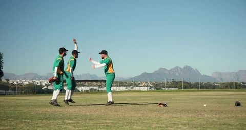 Diverse male cricket players celebrating leap on sunny suburban field with mountain backdrop