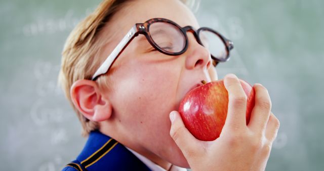 Boy wearing uniform and glasses, taking a bite of red apple in classroom. Ideal for educational websites, school promotional materials, healthy eating campaigns, and childhood development resources.