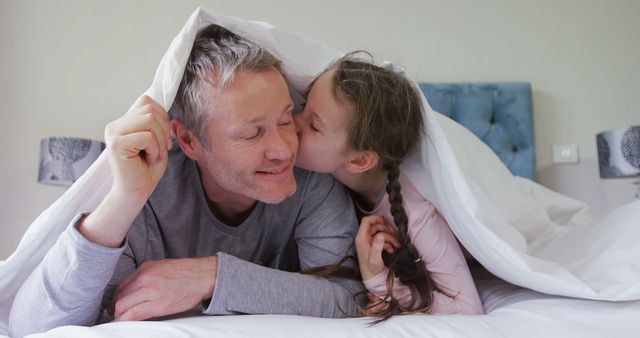 A Caucasian middle-aged man enjoys a playful moment as a young girl gives him a kiss on the cheek under a white blanket, with copy space. Their affectionate interaction captures a heartwarming family bond.