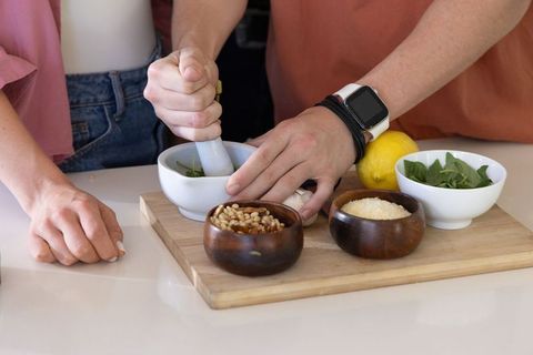 Couple preparing fresh pesto with mortar and pestle on wooden cutting board, home kitchen