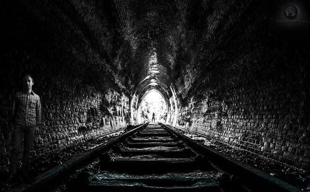 This image shows a man standing inside a dark, moody tunnel with light shining at the end. The rail tracks lead directly to the light, creating a strong focal point and a sense of perspective. This image can be utilized for conveying themes of hope, journey, mystery, solitude, and the human condition. Perfect for use in conceptual projects, storytelling visuals, background images, and exploration-related content.