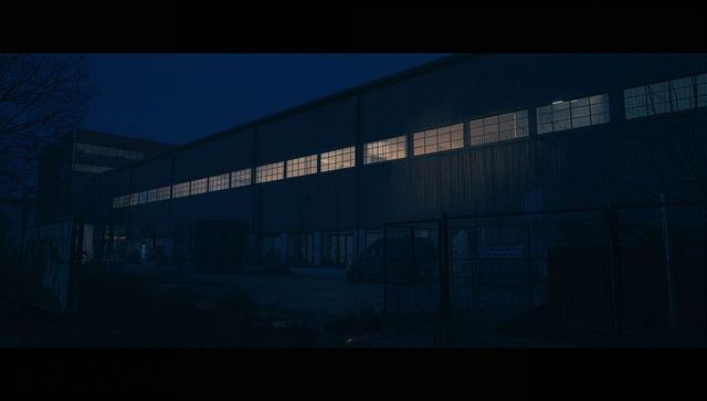 Industrial warehouse at night with glowing high windows and chain-link fence, corrugated metal