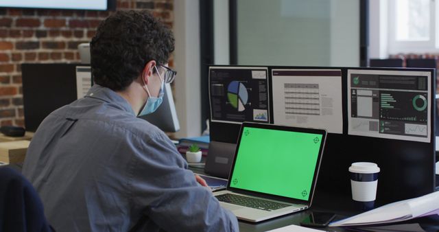 Person working at desk with multiple monitors displaying data and various charts. Laptop screen shows green placeholder. Wearing protective mask, indicating health safety measures during pandemic. Ideal for illustrating modern office environments, data analysis, or remote work setups.