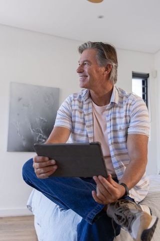 Mature man sitting on bed using tablet in minimalist bedroom, relaxed digital lifestyle