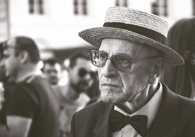 Elderly man wearing a straw hat and bow tie in outdoor setting with a crowd in background. His serious expression and classic outfit give vintage and timeless vibe. Perfect for themes like aging, vintage fashion, and societal commentary. Ideal for use in articles related to senior life, fashion history, or nostalgic moments.