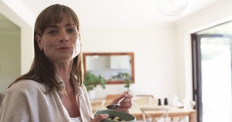 A middle-aged Caucasian woman is enjoying a healthy meal at home, filled with natural light. She appears relaxed and content in a modern dining room. Great for advertising healthy lifestyles, wellness promotion, and dining room design inspiration.