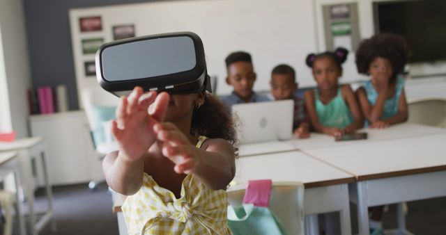 Group of young students engaging with virtual reality and technology in a modern classroom. The photo showcases a girl wearing a VR headset while other children in the background use a laptop, representing interactive, hands-on learning experiences in an educational environment. Ideal for illustrating articles and materials related to educational technology, modern education methods, and child development.