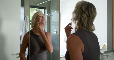 Mature woman applying jade facial roller at bathroom vanity mirror morning skincare