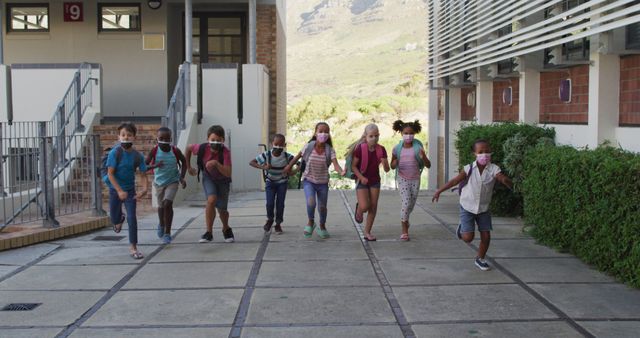 Diverse group of schoolchildren wearing backpacks and face masks running at school yard. children at primary school in summer during coronavirus covid 19 pandemic.