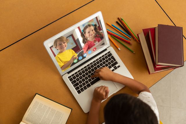 Child engaged in an online learning session with classmates through a video call on a laptop. The table is scattered with open books and colored pencils, indicating a study environment. Suitable for illustrating topics related to remote education, virtual classrooms, and the use of technology in schooling.