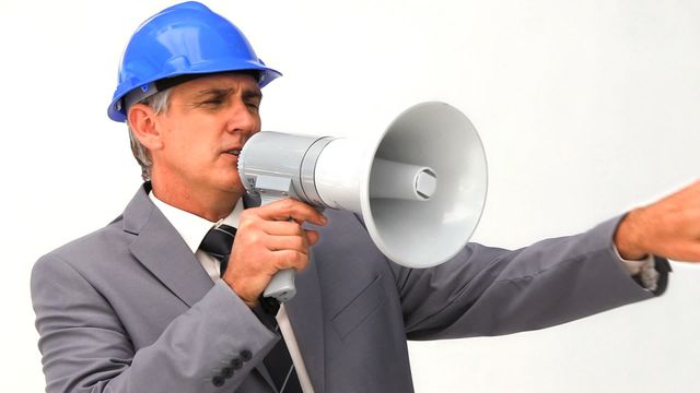 Professional mature businessman wearing a blue safety helmet and grey suit using megaphone for communication. Excellent for themes of leadership, authority, and industrial management. Useful in corporate, safety training materials, and promotional content about effective communication.