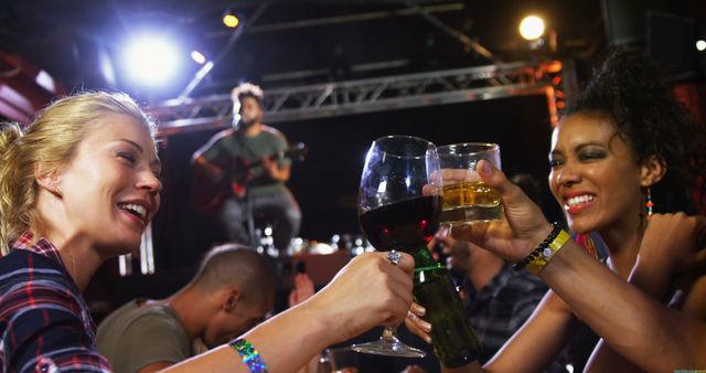 Two young women, one Caucasian and one African American, are toasting with drinks at a lively music event, with copy space. Their cheerful expressions and the ambient stage lighting capture the essence of a fun night out with friends.
