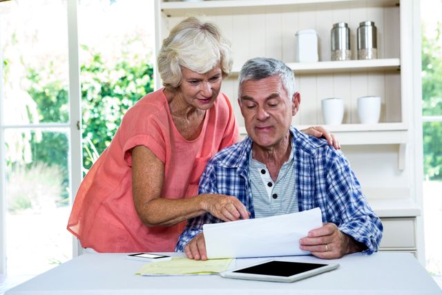 Senior couple sitting at kitchen table, reviewing documents together. The woman is pointing at the papers while the man is holding them. A tablet and other papers are on the table. This image is ideal for illustrating themes related to retirement planning, financial discussions, elderly lifestyle, and home life.