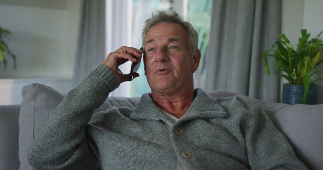 An older man is smiling while talking on the phone. He is dressed casually in a gray sweater and sitting on a comfortable couch. The room has a cozy ambiance with a plant and curtain in the background. This image may be used for themes related to communication, technology for seniors, happiness, and relaxation.