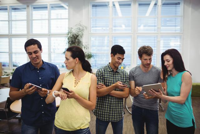 Group of diverse colleagues standing in office, using mobile phones and tablets. Perfect for depicting modern office environments, teamwork, and technology integration in business settings. Suitable for use in articles, presentations, and websites focused on business collaboration, technology use in the workplace, or modern work culture.