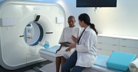 Diverse female patient and clinician discussing ct scan beside ct gantry using tablet