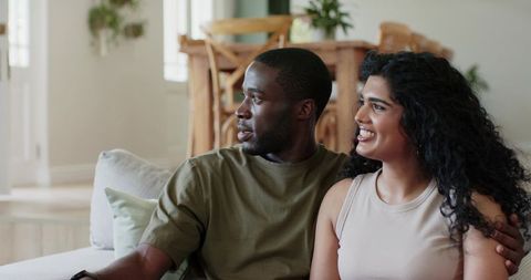 Diverse couple relaxing on light sofa in open-plan living room, smiling and looking left
