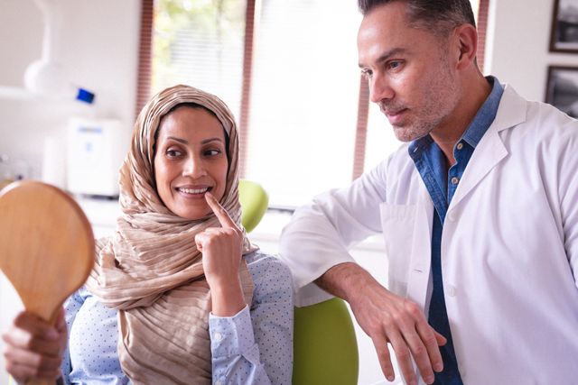 Female patient wearing hijab discussing dental health with male dentist in clinic. Patient holding mirror and pointing at teeth. Ideal for use in healthcare, dental care, and medical consultation contexts.