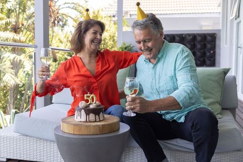 Senior couple celebrating 50th milestone on sunlit patio with wine and chocolate cake