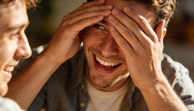 Young man laughing while holding head during cozy indoor friends hangout, candid smile