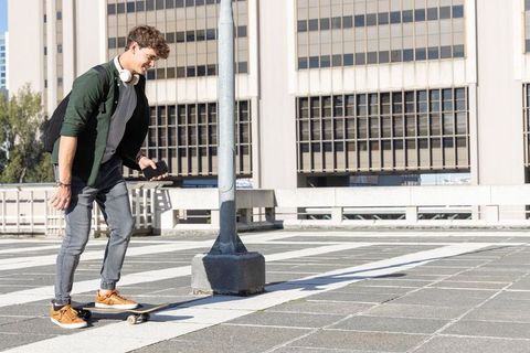 Skateboarding young man checking phone on urban rooftop parking deck with backpack