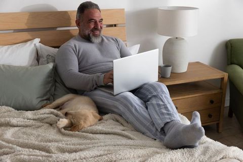 Senior man working on laptop in cozy bed with sleeping puppy, pajama lounge and remote work comfort