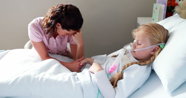 Mother sitting beside young daughter in hospital bed who is wearing oxygen mask, holding her hand. Mother showing concern and comforting sick child. Suitable for themes related to pediatric health care, family support in medical settings, childhood illness, and hospital care.