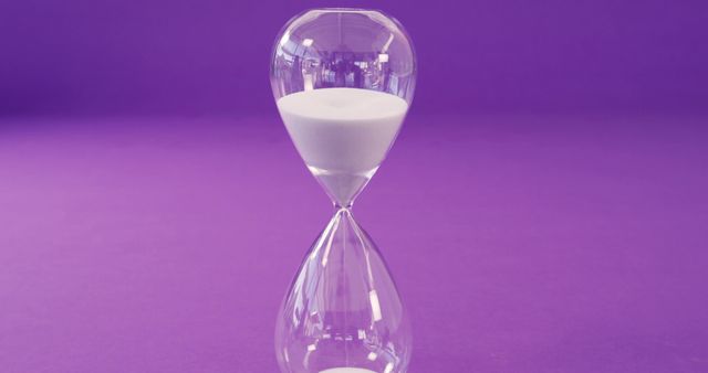 Elegant clear hourglass displayed with white sand flowing against rich purple background. Symbolizing time passage, deadlines, and punctuality. Ideal for business presentations, motivational content, time management subjects, and minimalistic designs.
