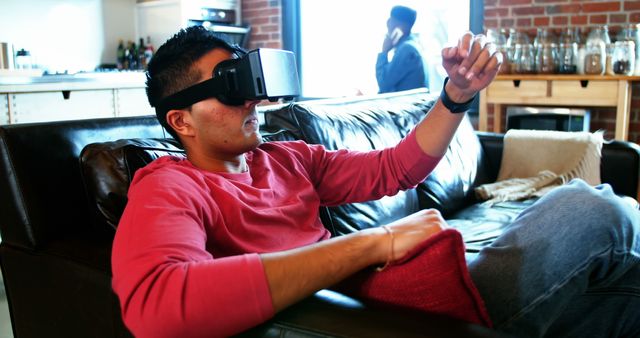 Man relaxing on couch inside modern living room while using virtual reality headset. Perfect for illustrating technology use, virtual gaming, modern lifestyle, and relaxation at home. Ideal for articles on VR advancements, immersive experiences, or technological relaxation.