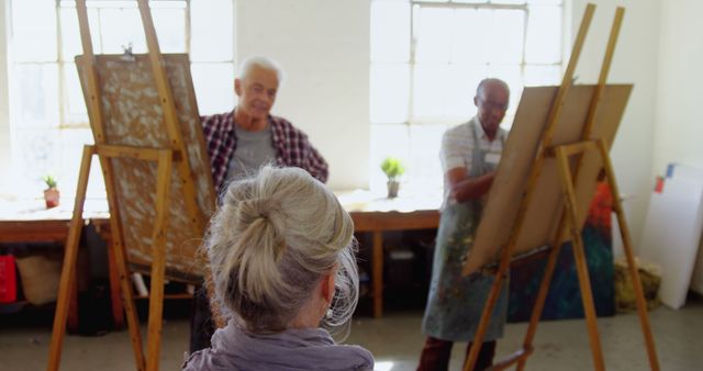 Senior artists standing at easels painting in an art studio with natural light. Suitable for content related to senior activities, art classes, creativity, hobbies, and retirement life.