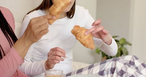 Two friends sharing croissants in checkered pajamas, sipping morning drink on couch