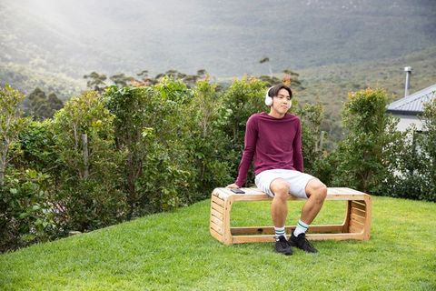 Young Asian man sitting on wooden bench listening to music with headphones in serene garden