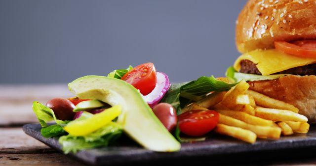 A delicious cheeseburger with lettuce and tomato is paired with a side of golden fries and a fresh salad garnished with avocado slices, offering a tempting mix of indulgence and freshness. This meal presents a classic fast-food option with a side of vegetables for a balanced approach to dining.