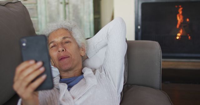 Senior woman lying on couch and using smartphone, with lit fireplace in background. Perfect for topics related to senior lifestyle, technology use among elderly, home comfort, relaxation, and digital connectivity for older adults.