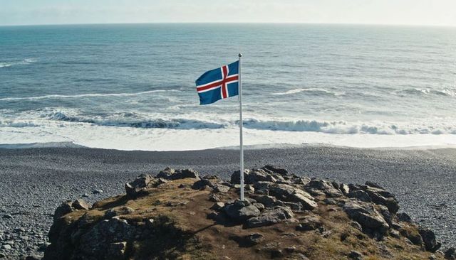 Icelandic flag fluttering on rocky headland over pebble beach and atlantic waves