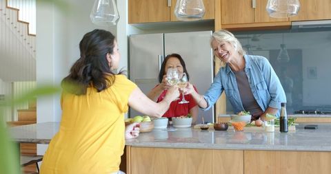 Women clinking white wine glasses over kitchen island while sharing salad and conversation