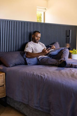 Mature african man reclining on bed using smartphone in modern minimalist bedroom