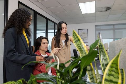 Multicultural team collaborating at open-plan desk pointing at monitor and tablet with office plants