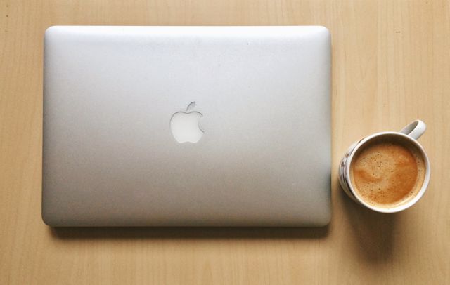 Overhead view of closed MacBook and cup of coffee on light wooden desk. Ideal for depicting workspace, tech productivity, work-from-home concepts, or coffee break during work.