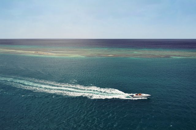 A speedboat cruises on a sparkling blue ocean under a clear sky. The aerial view captures the wake behind the boat, creating a dynamic sense of motion and adventure. Ideal for travel brochures, tourism websites, and water sport promotions suggesting leisure and fun during summer vacations.
