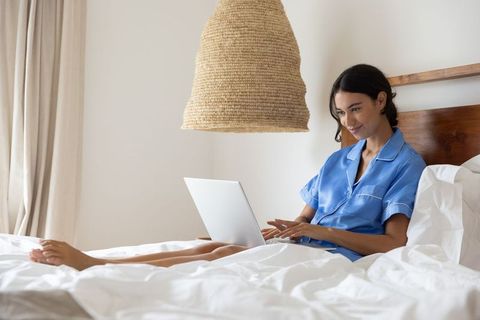 Woman in blue pajamas working on laptop in cozy minimalist bedroom with woven pendant lamp